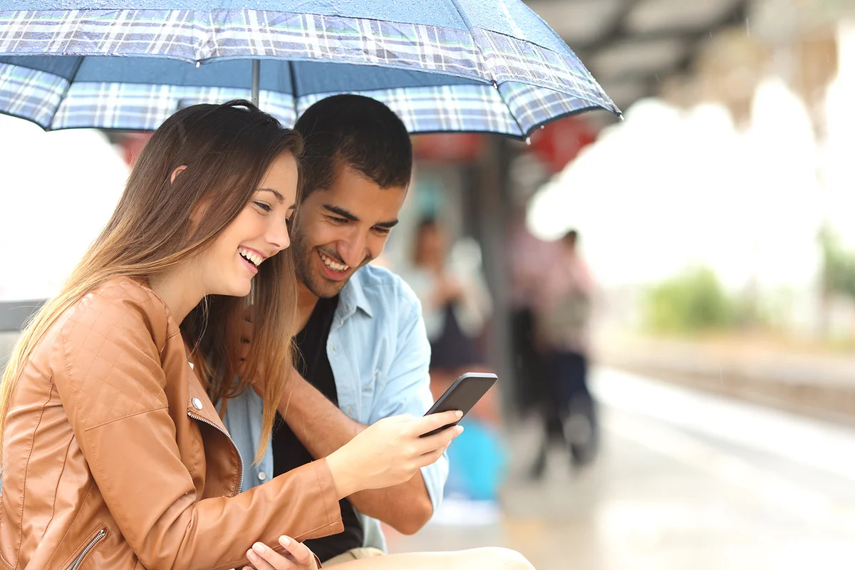 couple sharing a phone in a train station while wait under an umbrella in a rainy day