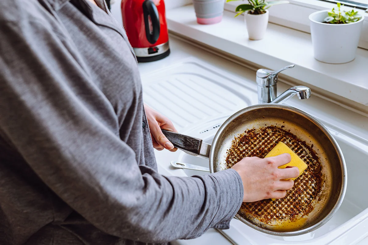 woman's hand washes burnt greasy frying pan with kitchen washcloth in sink