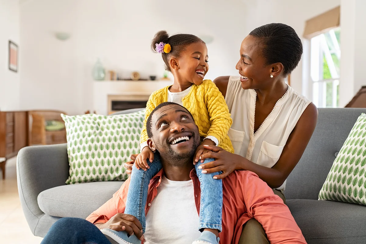 Cheerful little girl sitting on father shoulder while playing with mother at home