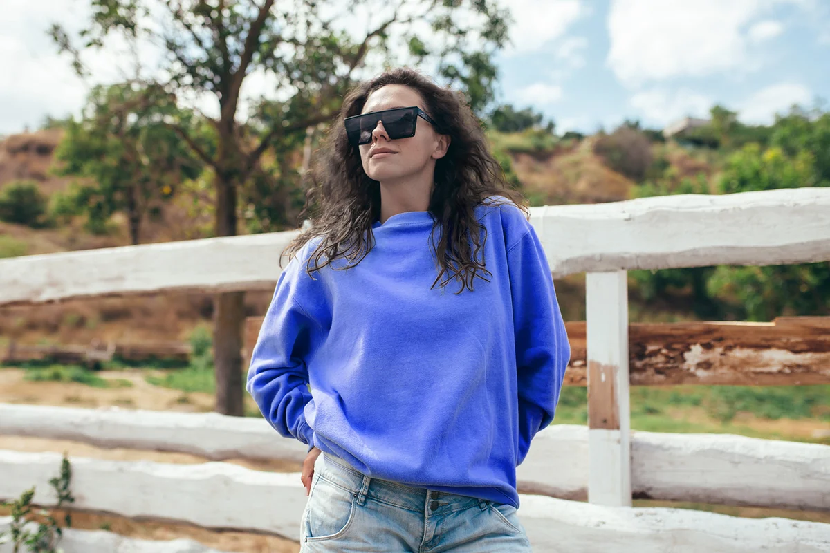 Beautiful young adult woman in blue blank sweatshirt and sunglasses on the background of a wooden fence in nature.