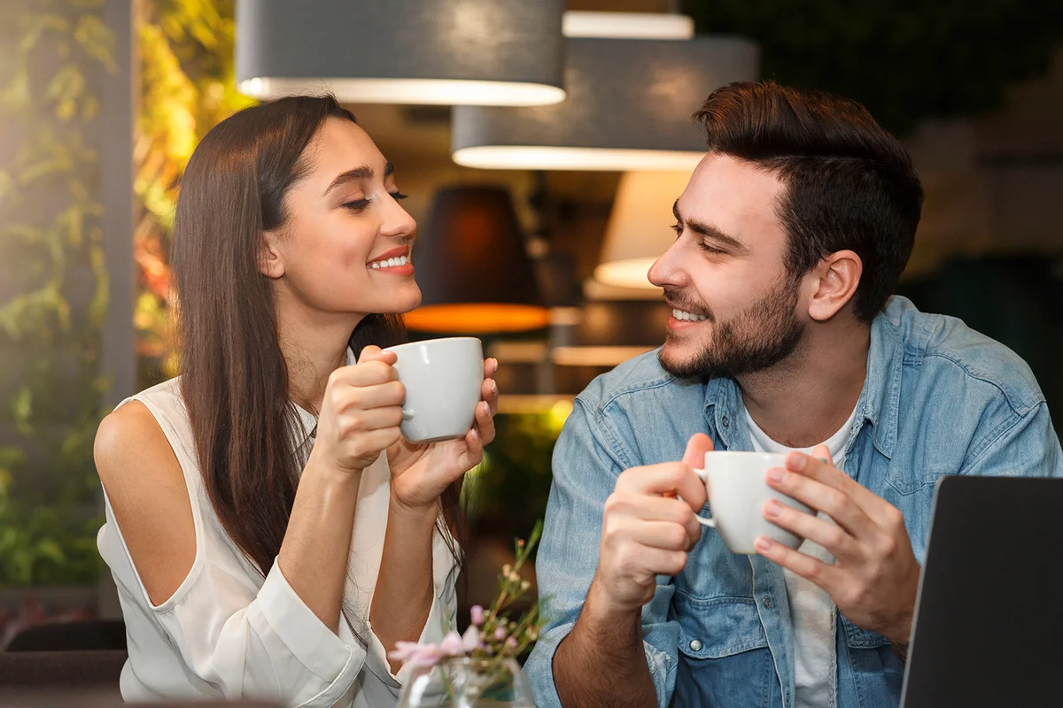 Young couple tasting coffee drinks enjoying flirt and conversation during weekend date sitting in cozy cafe