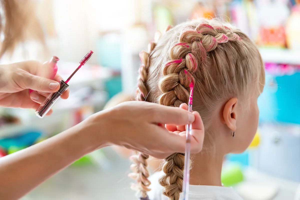Close-up hand holding brush make dyed braids with pink color on little daughter kid head.