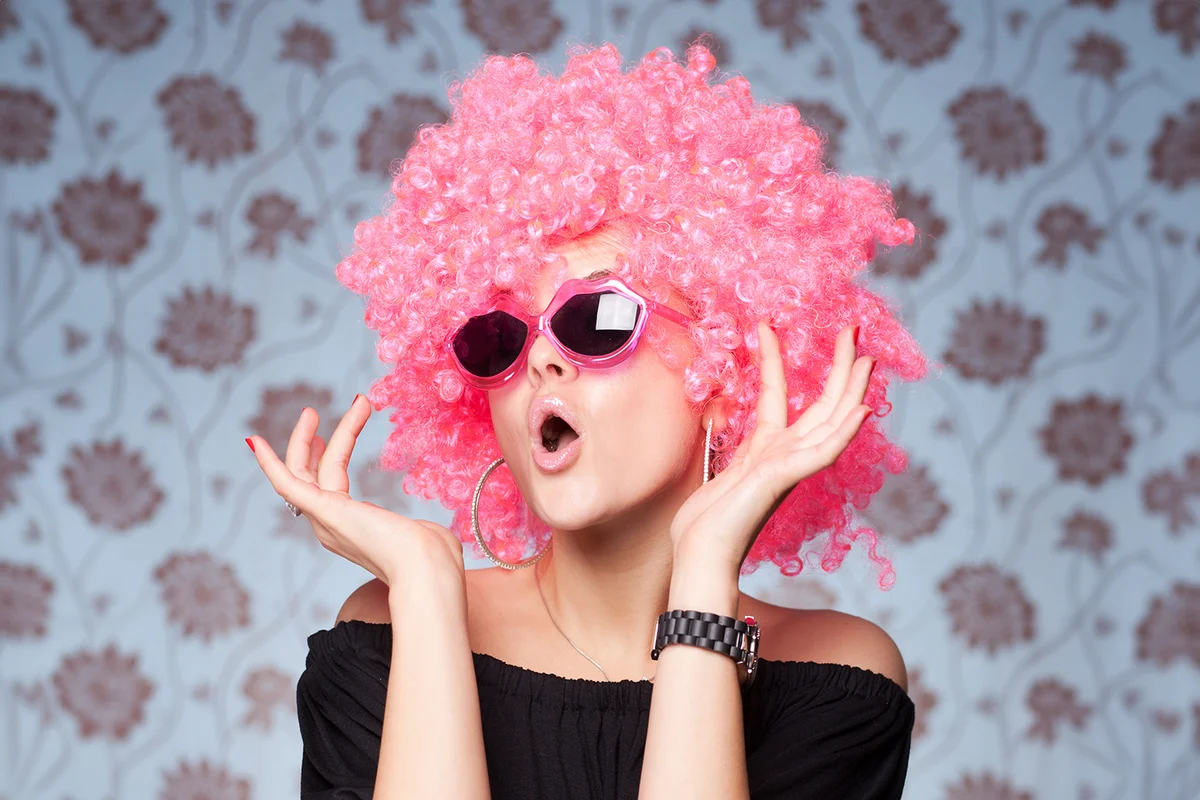 Funny girl in pink wig and glasses posing for camera across wallpapers background