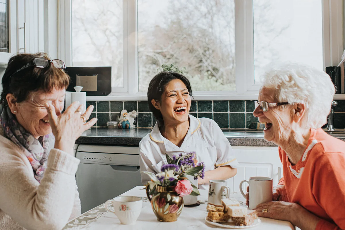 A carer laughs during a conversation with two older ladies at a kitchen table