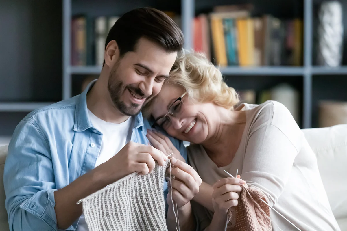 mom and adult son relax on couch in living room knitting together