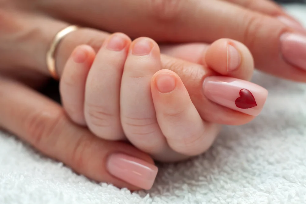 Close-up of a woman's hand with a heart design on her nail, being held by a baby's hand.