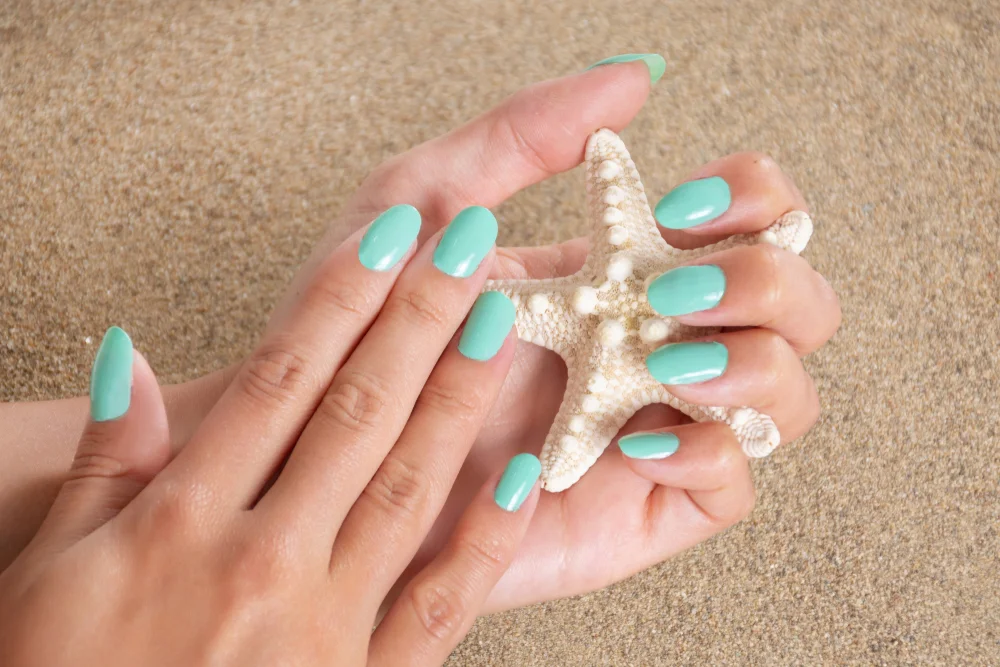 Turquoise color manicured hands holding a starfish with a sand background.
