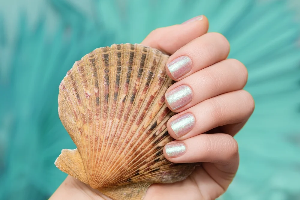 A female hand with a glittery pearl manicure holding a seashell on a turquoise background