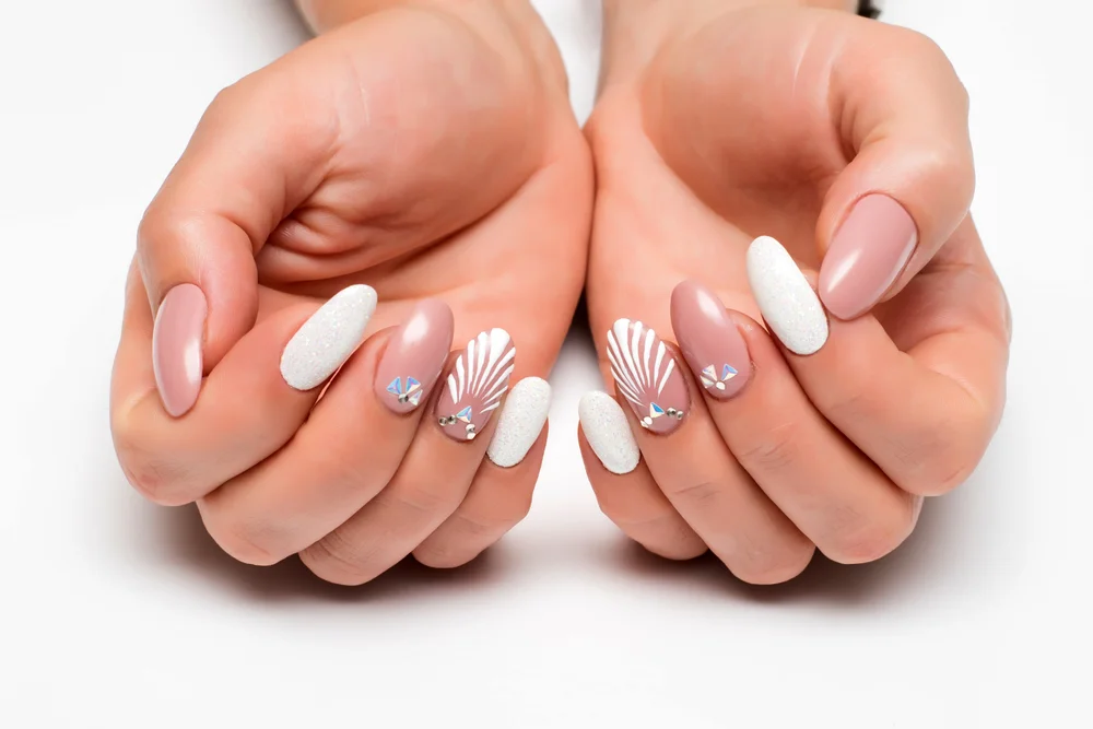 Hands with a nude and white manicure, embellished with shell drawings and crystals.