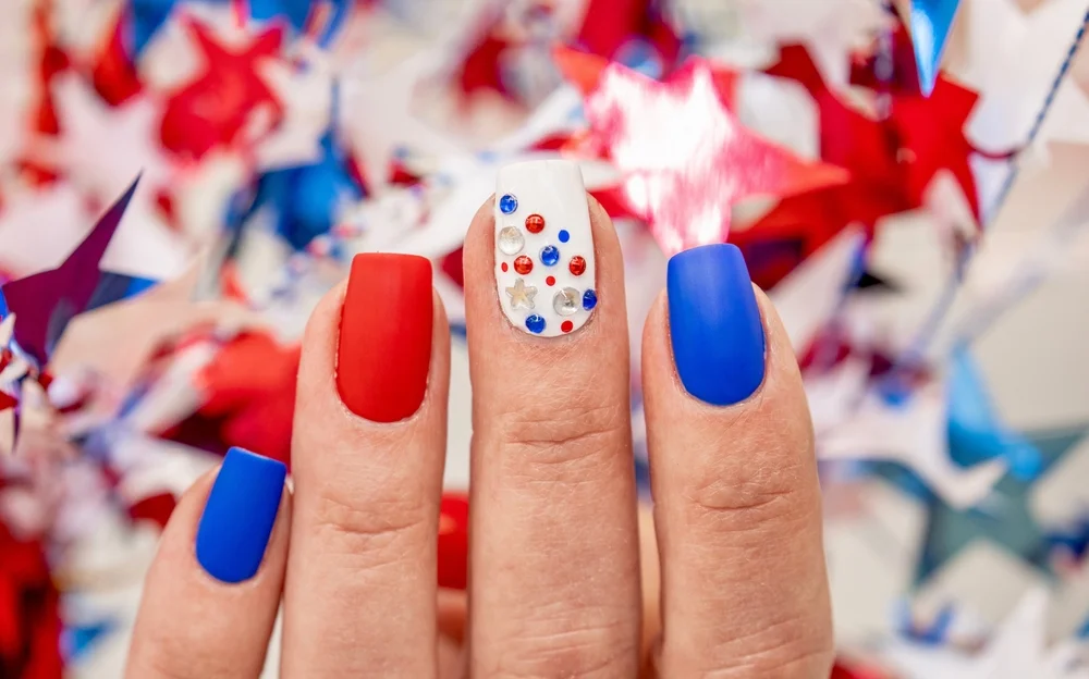 Close-up of a matte blue, red, and white manicure with embellishments and a festive US-themed background.