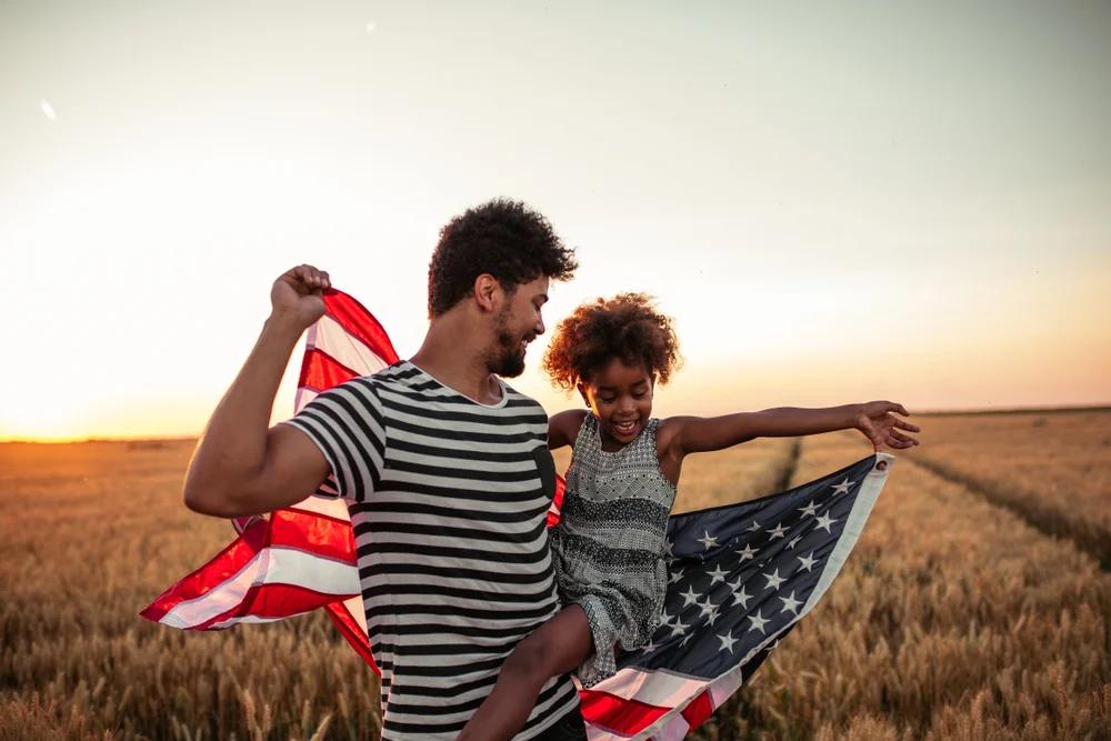 Candid medium shot of a father and his daughter holding the US flag in nature during a beautiful sunset.