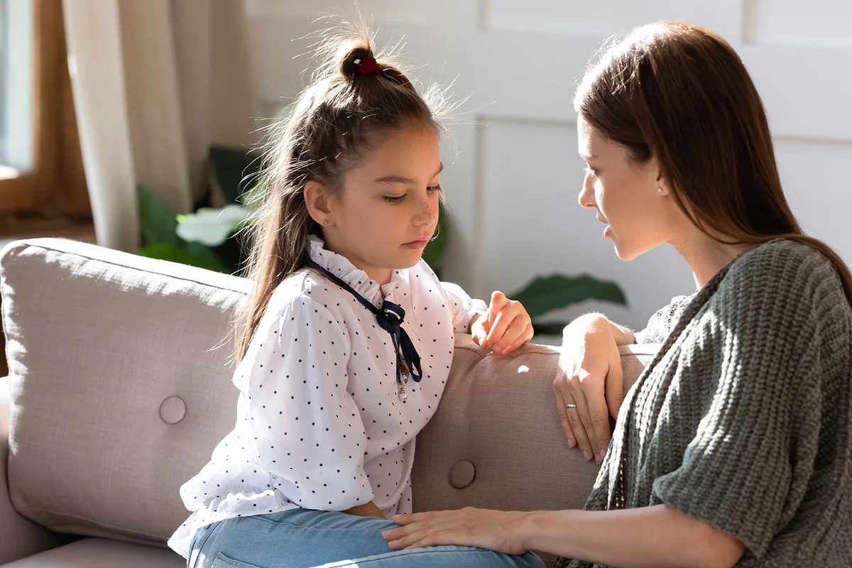 Upset small schoolgirl having trustful conversation with compassionate young mother, sitting together on sofa.