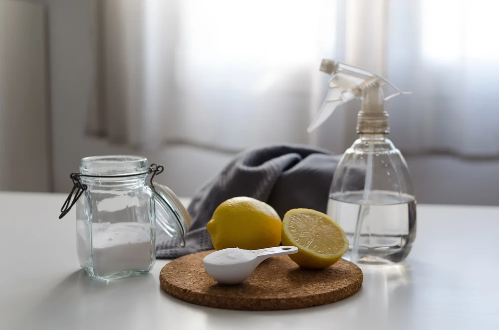 Eco spray bottle filled with water and vinegar. A cleaning cloth, soda, and lemons on a table against a bright window.