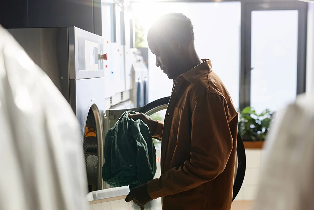 Side view of young black man standing by the washing machine putting clothes in a plastic container