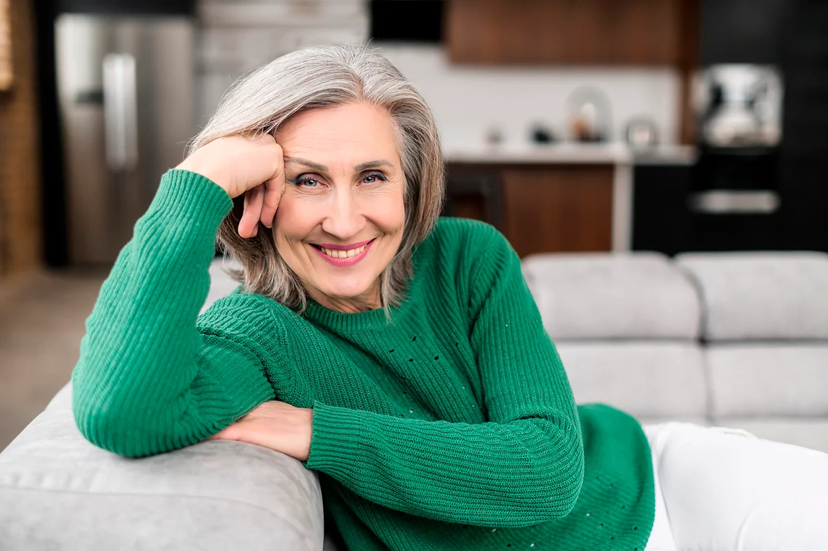 Portrait of charming senior woman with grey hair, smiling mature female sits on the couch at ease and looks at the camera. Beautiful carefree middle-aged