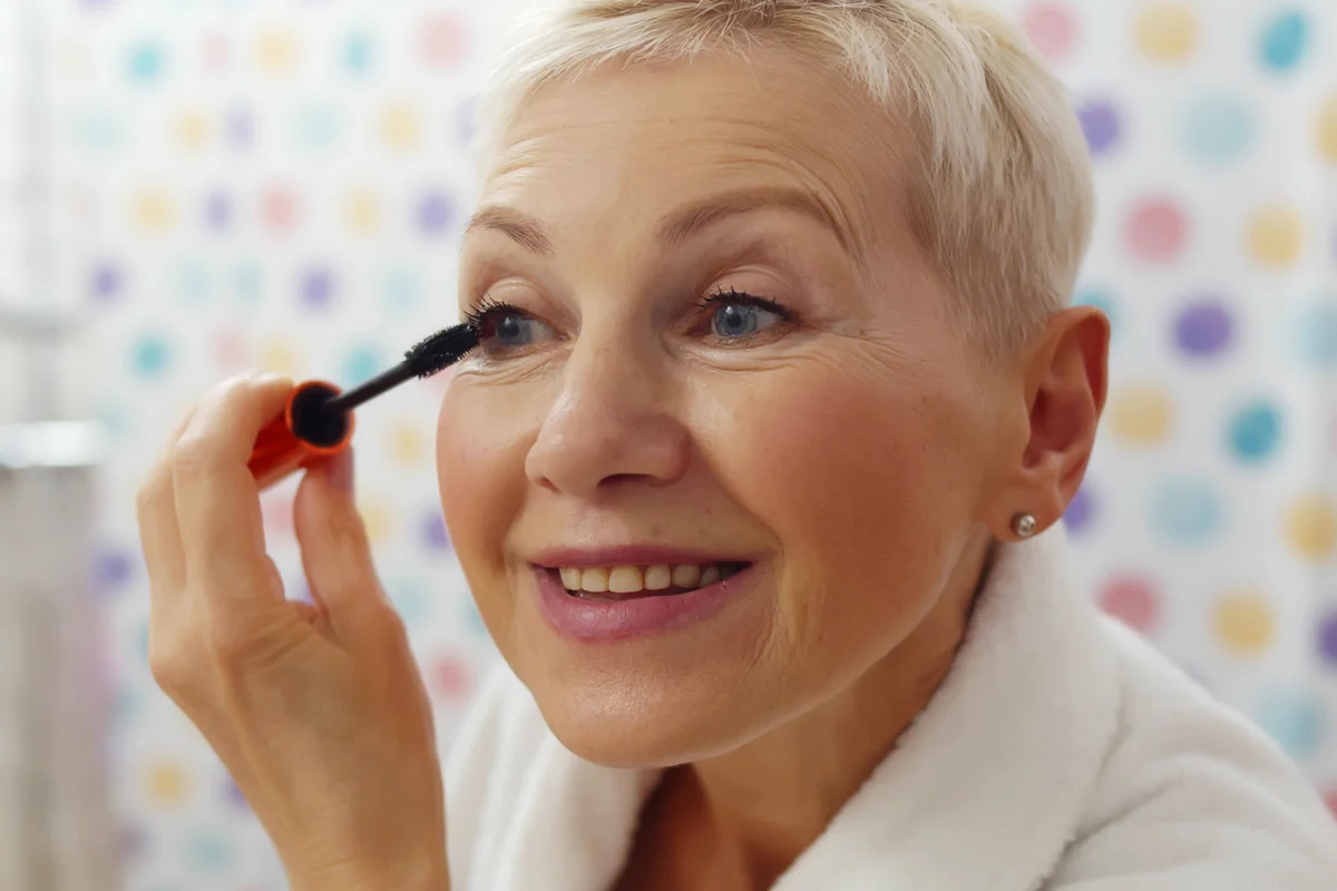 Mature woman putting mascara looking in mirror getting ready in morning
