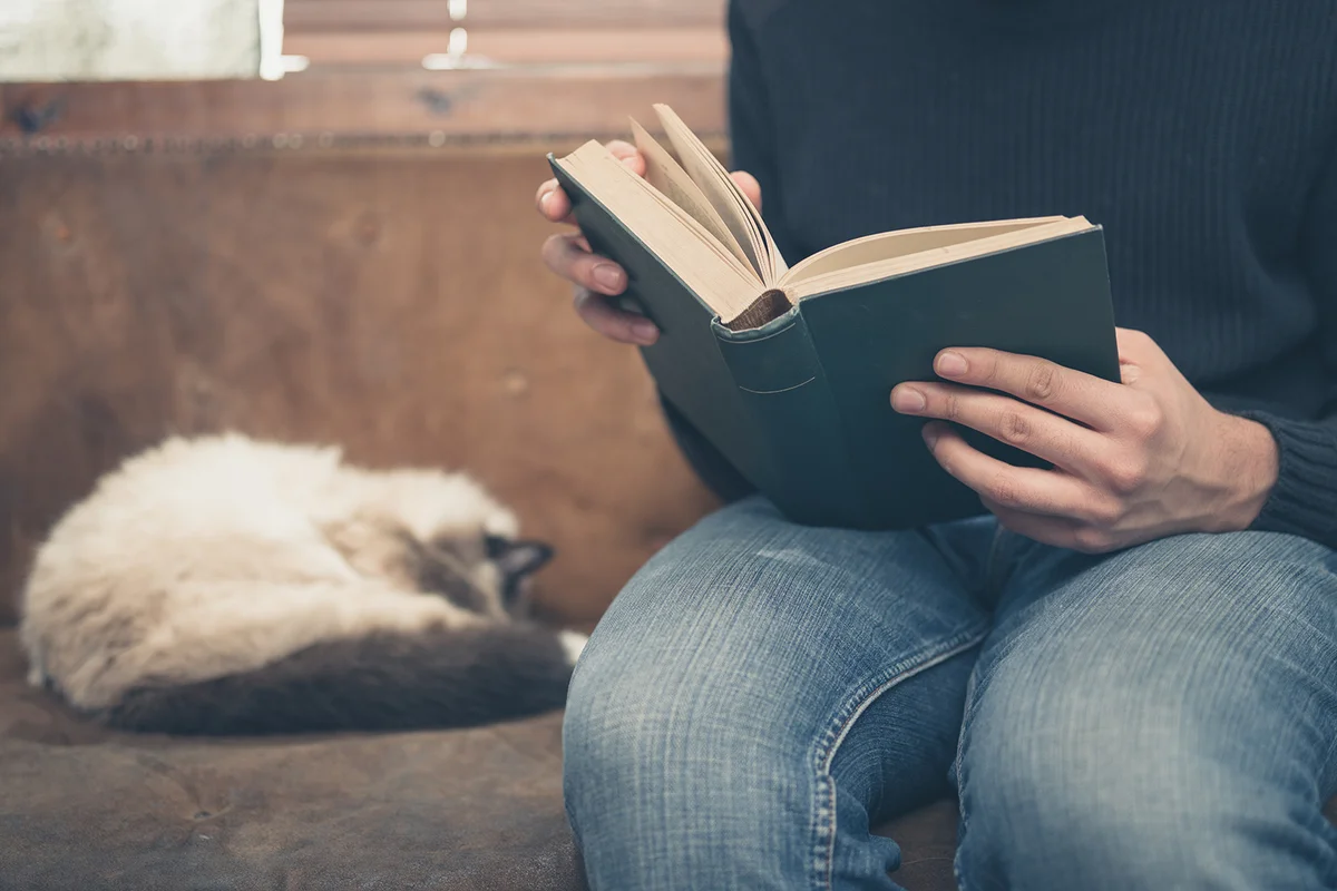 A young man is sitting on a sofa with a cat and is reading the Bible