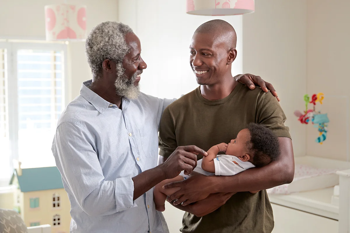Proud Grandfather With Adult Son Cuddling Baby Grandson In Nursery At Home