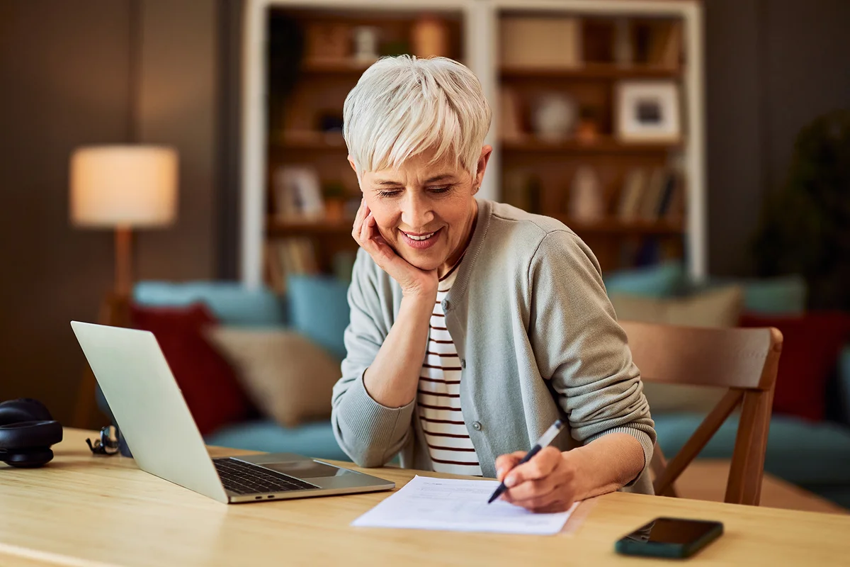 A happy senior adult female accountant with short hair working from home and writing financial reports in front of a laptop.