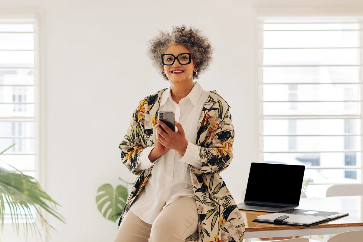 Happy senior businesswoman smiling at the camera while holding a smartphone in a boardroom