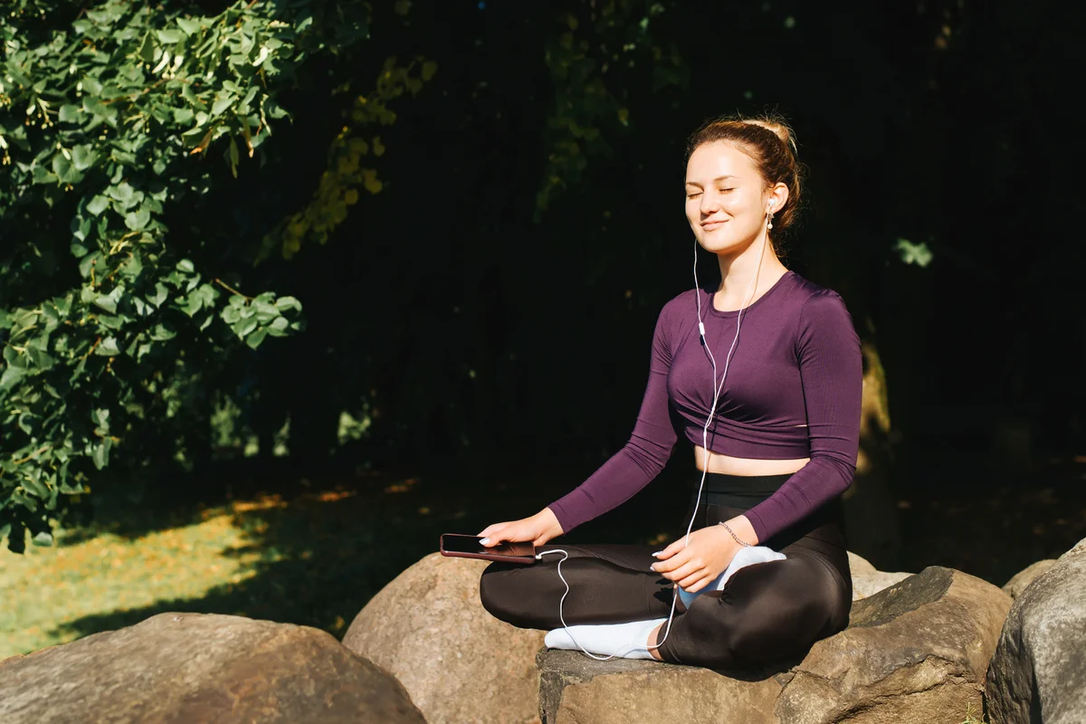 Smiling young woman in sportswear listens music on headphones using smartphone while meditating