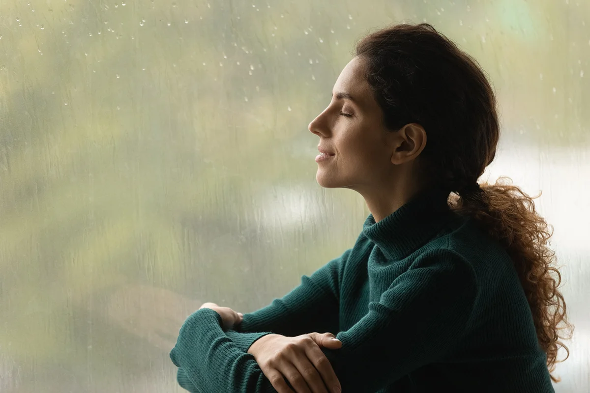 Side shot of serene latin woman relax by window with closed eyes listen sound of raindrops feel pleasure.