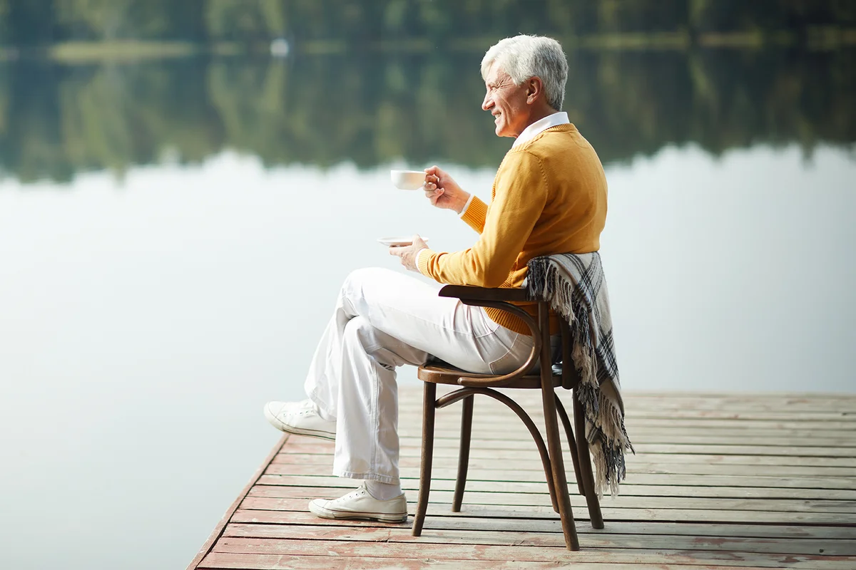 Happy relaxed handsome senior man wearing stylish clothing sitting on old chair with blanket on pier