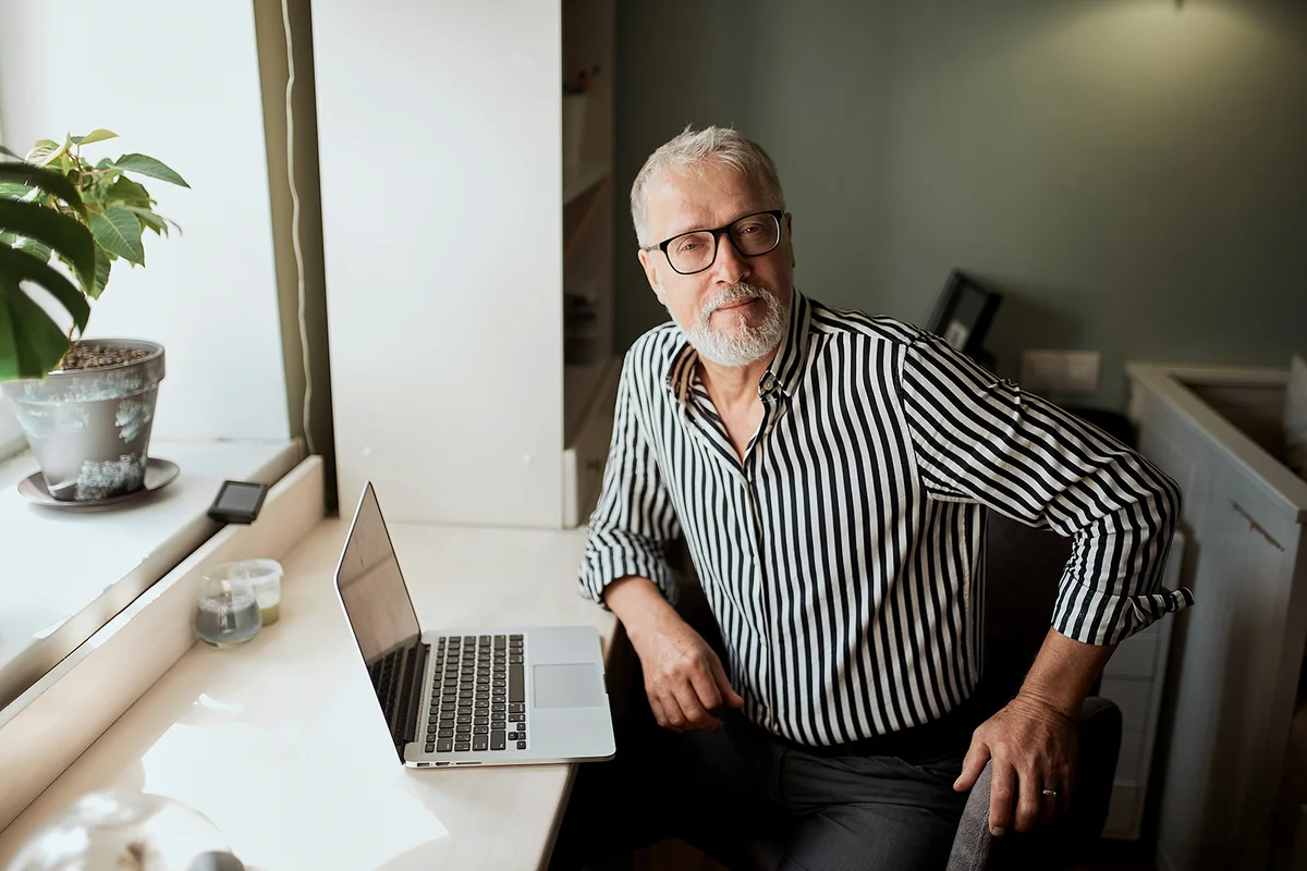 Trendy mature bearded man working from home with laptop. sitting at desk near window
