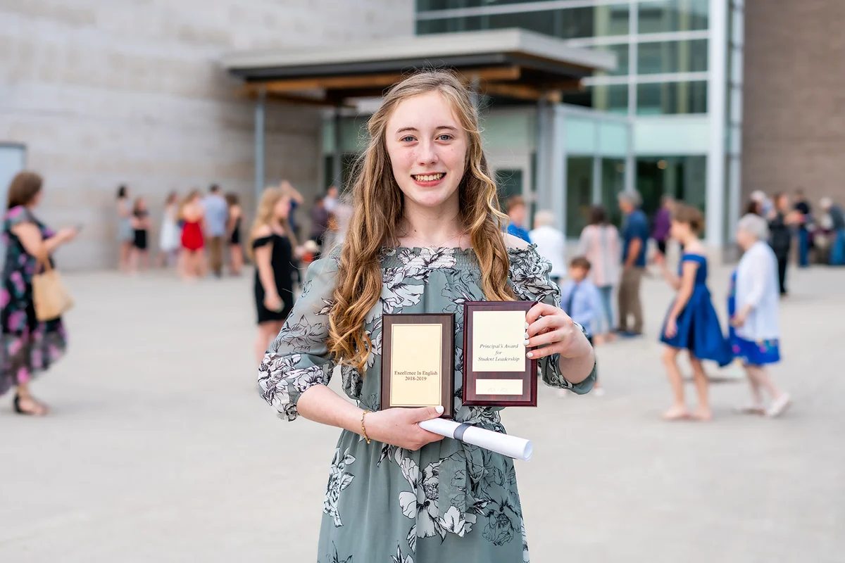 student standing in front of school with awards and diploma after Grade 8/Middle school graduation ceremony