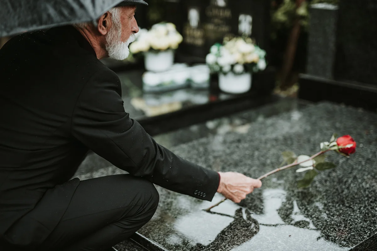 Elegant sad elderly man standing on the rain with umbrella and grieves at the grave of a loved person