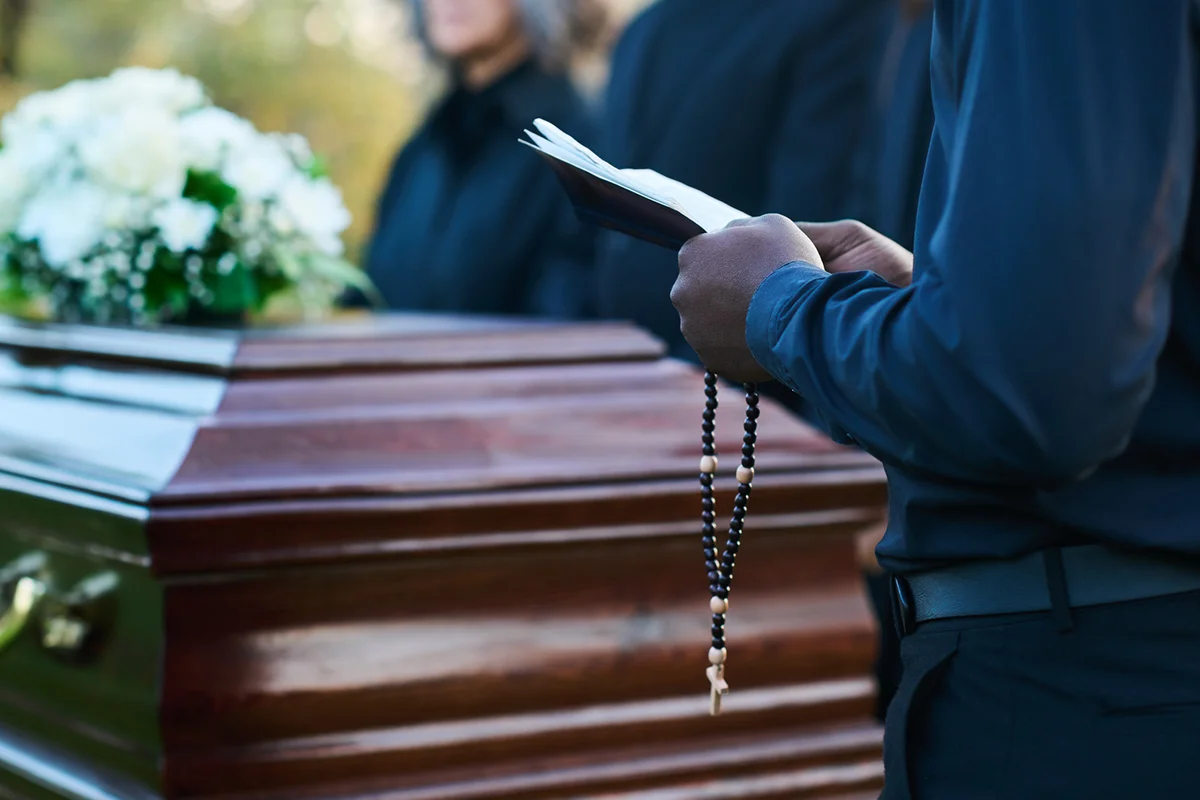 priest with open Holy Bible and rosary standing in front of coffin