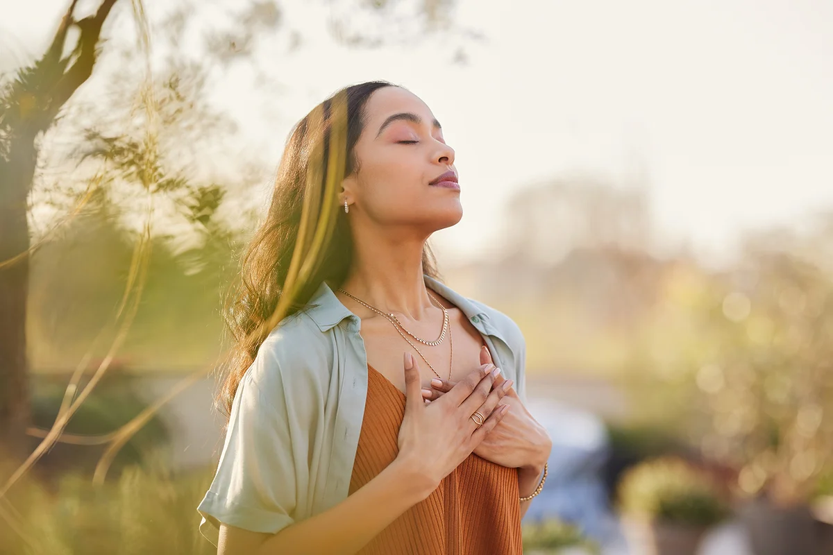 Young latin woman with hand on chest breathing in fresh air in a beautiful garden during sunset.