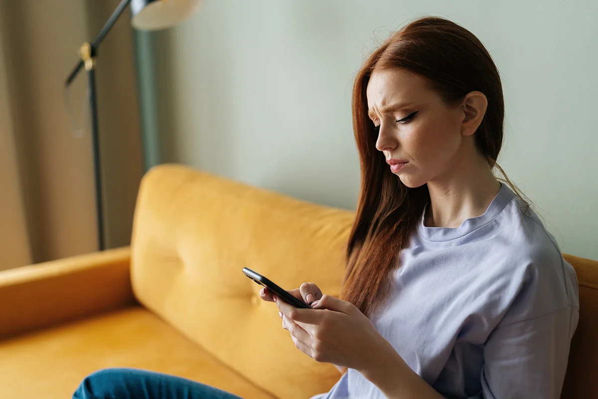 Sad red-haired young woman looking at smartphone
