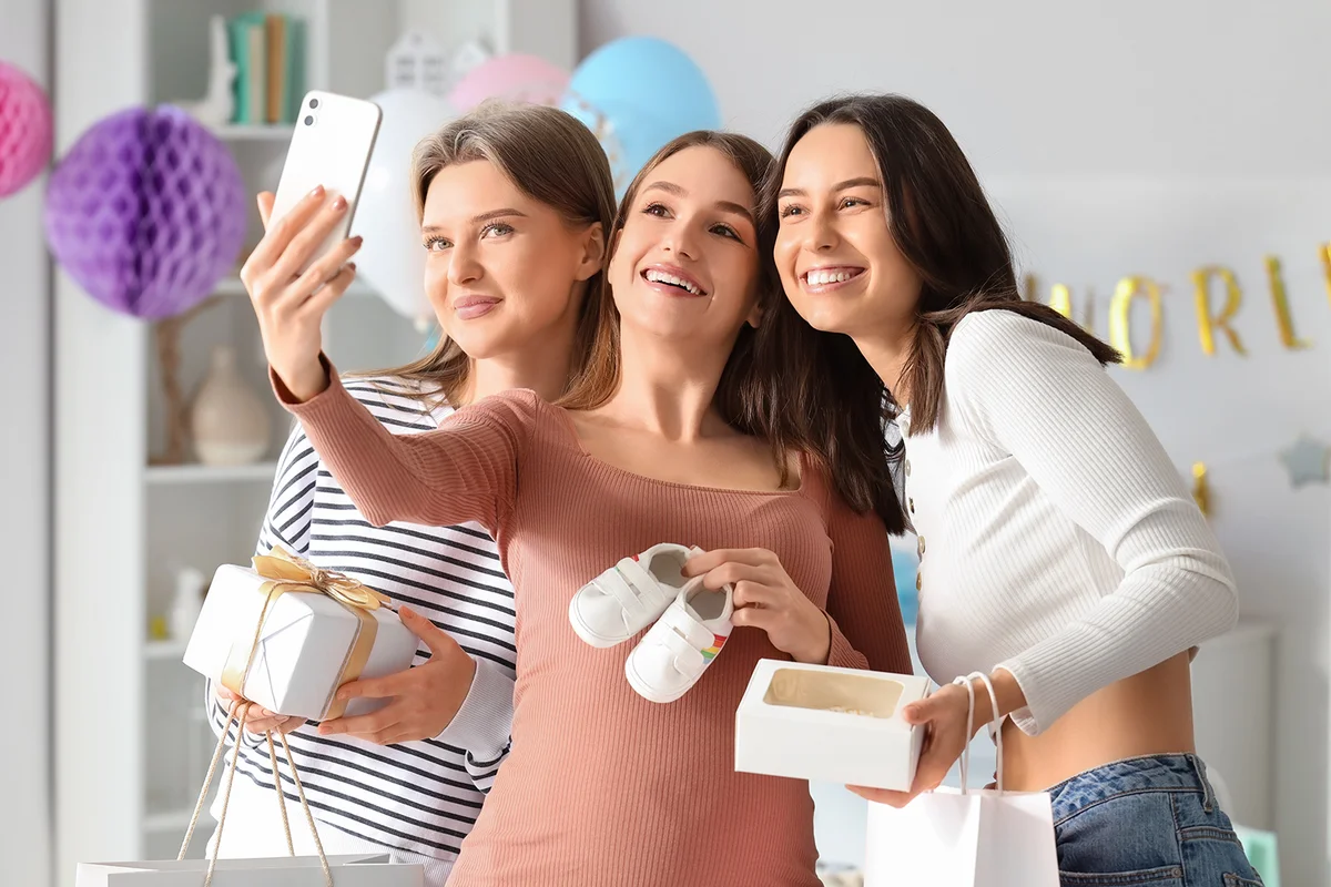 Young pregnant woman with her friends and gifts taking selfie at baby shower party