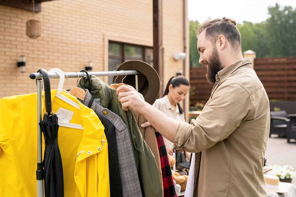 Smiling bearded man in shirt standing at rack and choosing clothes at yard sale