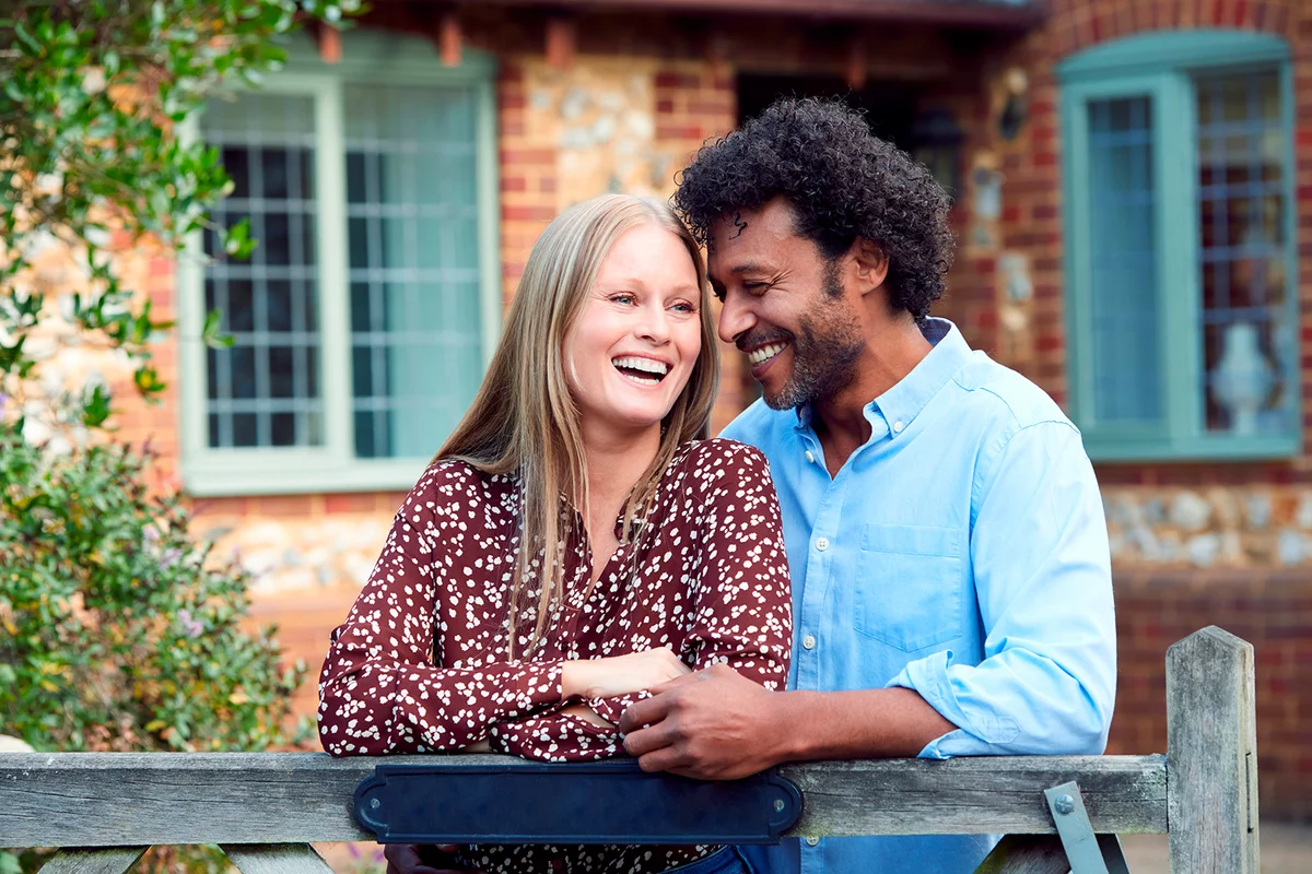 Portrait Of Loving Couple By Gate Holding House Keys Outside New Home In Countryside