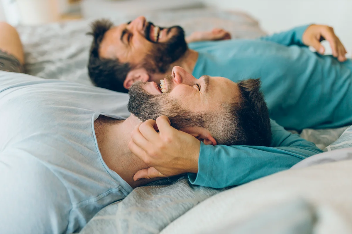 Happy gay couple lying down on the bed at home, hugging and flirting.