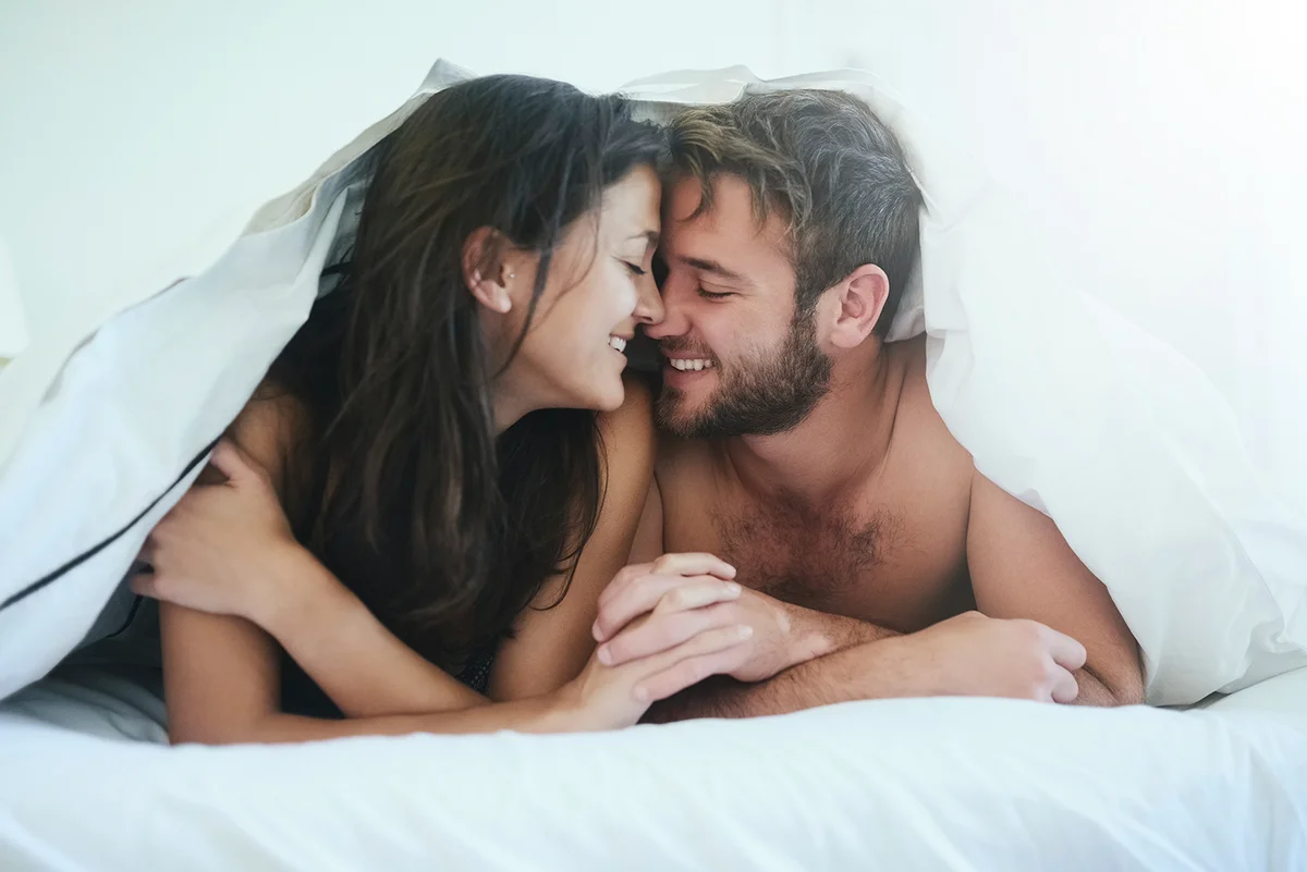 Love, cover and couple in bed with kiss, romance and happy morning bonding in apartment. Relationship, man and woman in bedroom with intimate embrace, passion and relax with care on weekend in home