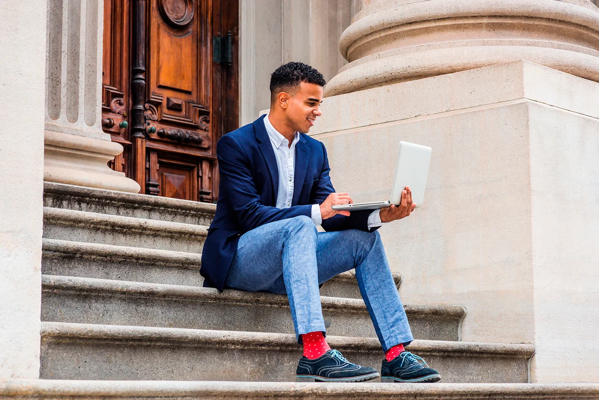 African American college student studying in New York, wearing blazer, gray pants, red socks, sneakers, sitting on stairs outside office building, working on laptop computer. Color filtered effect