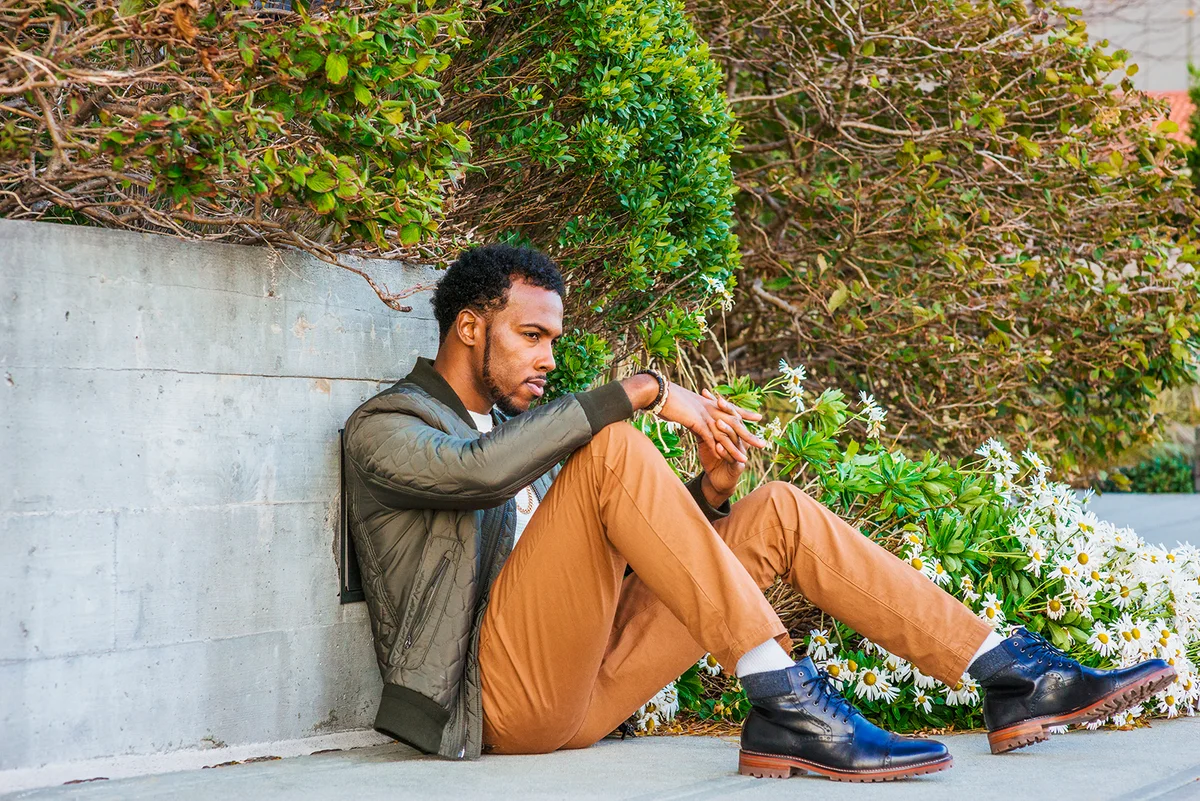 Winter Street Fashion in New York. African American guy wearing green coat, white knitting under sweater, brown pants, black leather boot shoots, sits against wall, sad, thinking. Filtered effect.