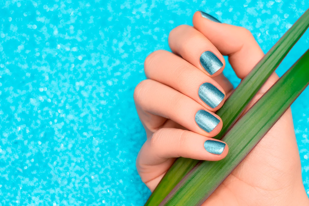 Female hand with light blue nail design. Glitter blue nail polish manicure. Female model hand with green plant leaves on glitter blue backround. Copy space.
