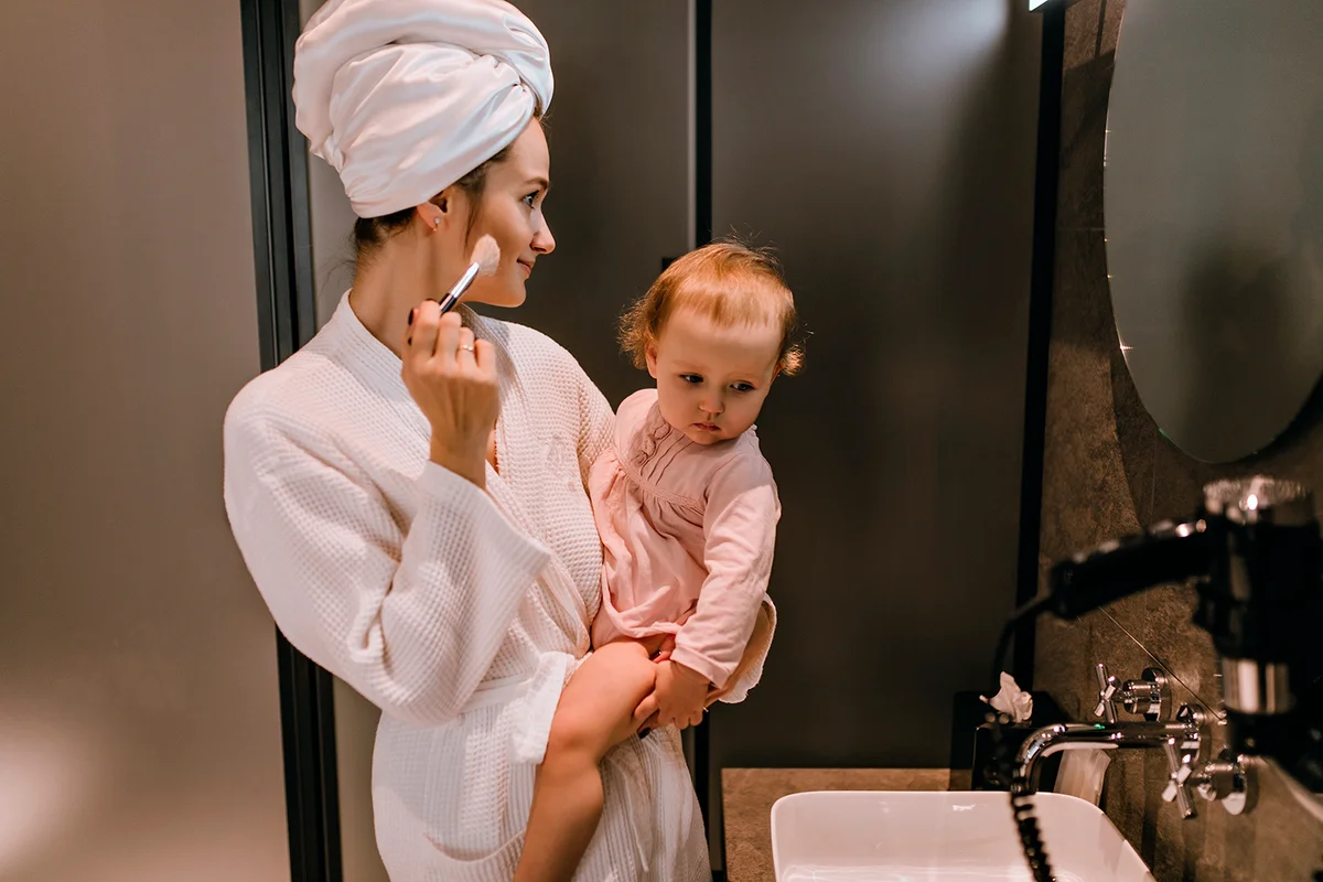 Young woman in bathrobe holds blonde daughter and does her own makeup, looking in bathroom mirror