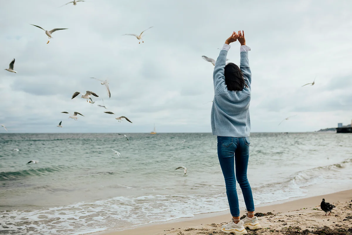 Beautiful girl on a background of azure sea. Photo from the back. Girl looking at the calm sea and flying seagulls on the background. Hands up, freedom, calm. Copy space.