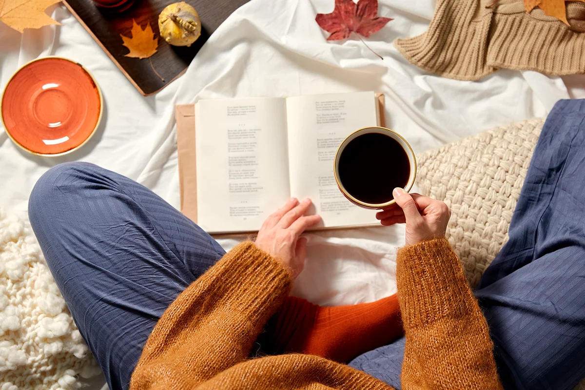 people, season and leisure concept - young woman drinking black coffee and reading book at home in autumn