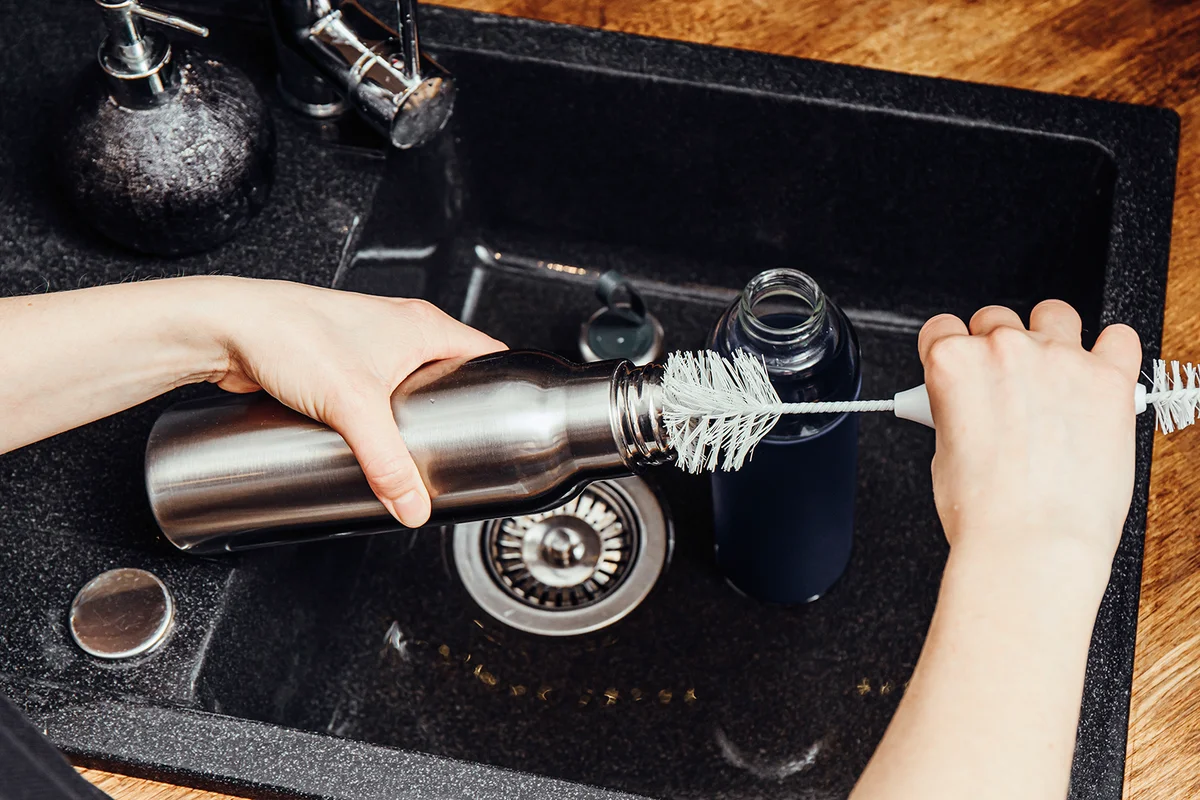 Washing cleaning reusable drinking water bottle with bottle cleaning brush in home kitchen. Sustainable lifestyle concept.