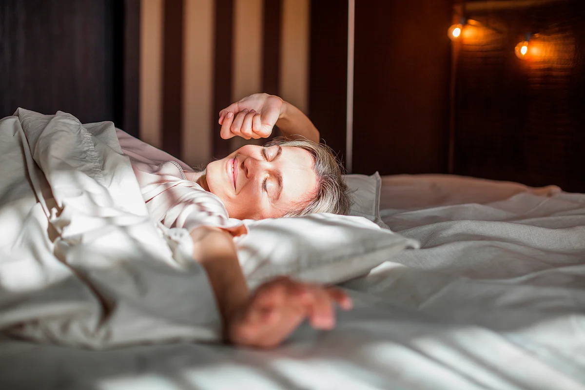 Good morning. Middle aged beautiful relaxed female with closed eyes in pajamas waking up in her bed fully rested, happy woman stretching after night sleep