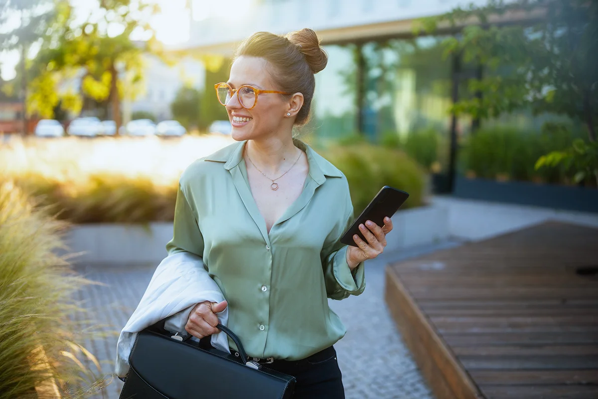 smiling modern woman worker near business center in green blouse and eyeglasses with briefcase using smartphone.