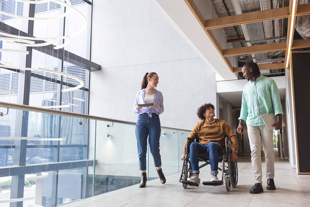 Diverse group of friends walking and talking in modern hallway in a modern business office. African American man in green shirt, biracial man in wheelchair, Caucasian woman in blue jeans, unaltered