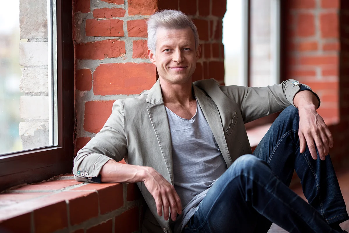 Casual mature man sitting by window with brick wall, relaxed pose, hand to head. Natural light ambiance.