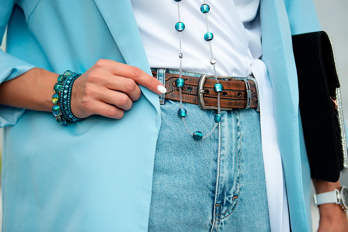Fashionable women's accessories. Detail of the body of a model dressed in light blue high-waisted jeans, an ornamented leather belt and a blue jacket. Jewelry from blue stone beads and bracelet.